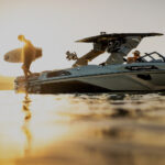 A person stands on the back of a Centurion boat holding a surfboard at sunset, while another person sits at the helm.