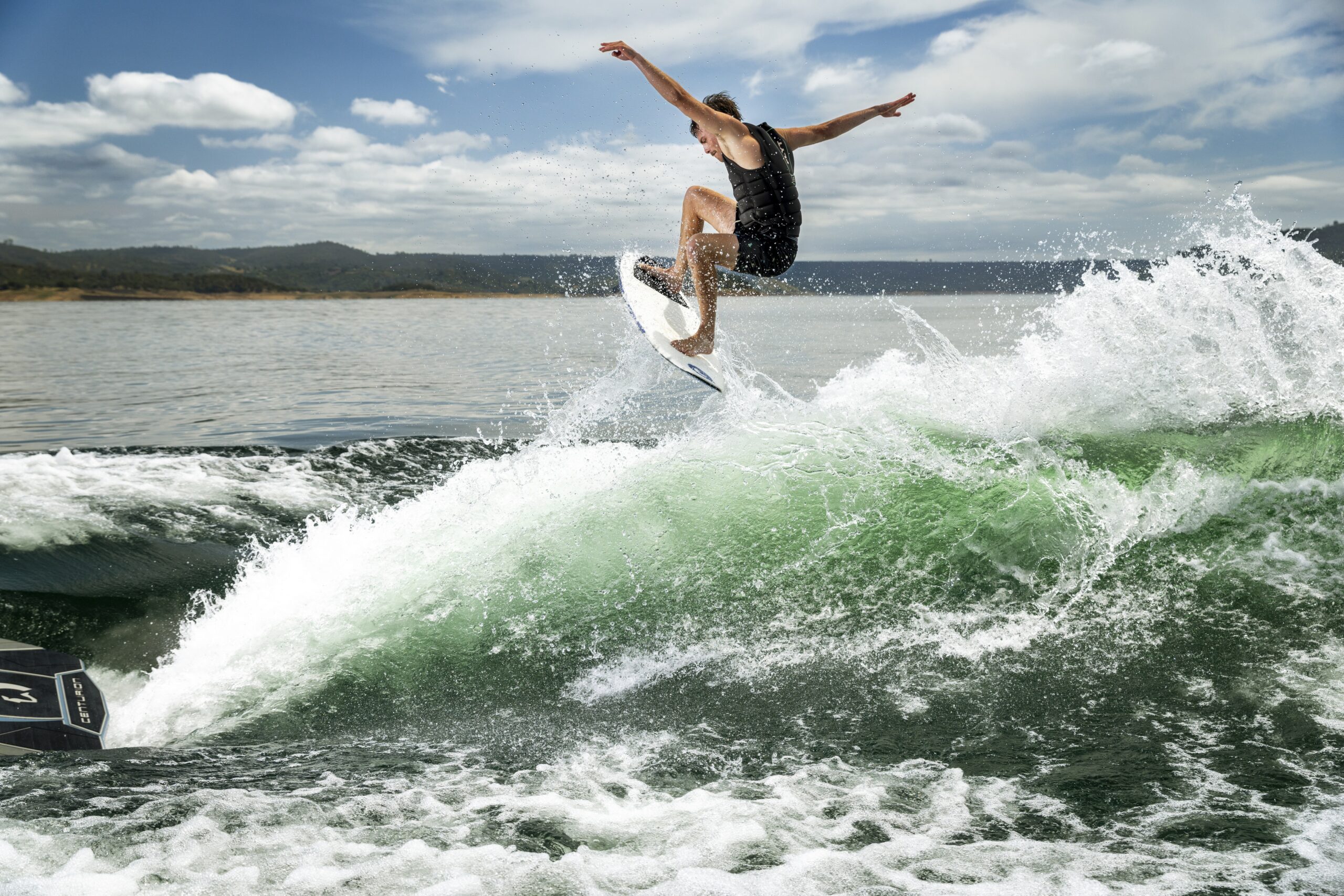A person wearing a black outfit rides a surfboard and jumps over a wave on a sunny day, with water splashing around and distant hills in the background.