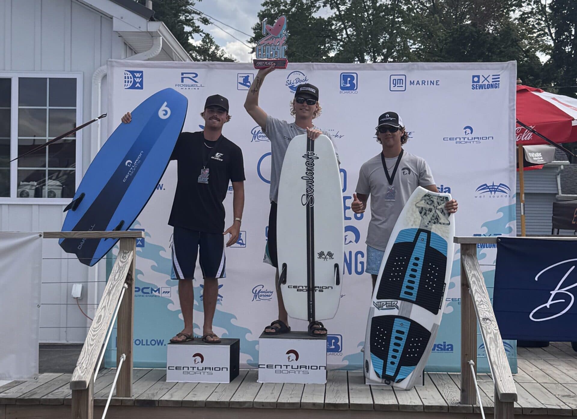 Three surfers stand on a podium holding boards, posing for a photo at a surfing competition award ceremony outdoors.