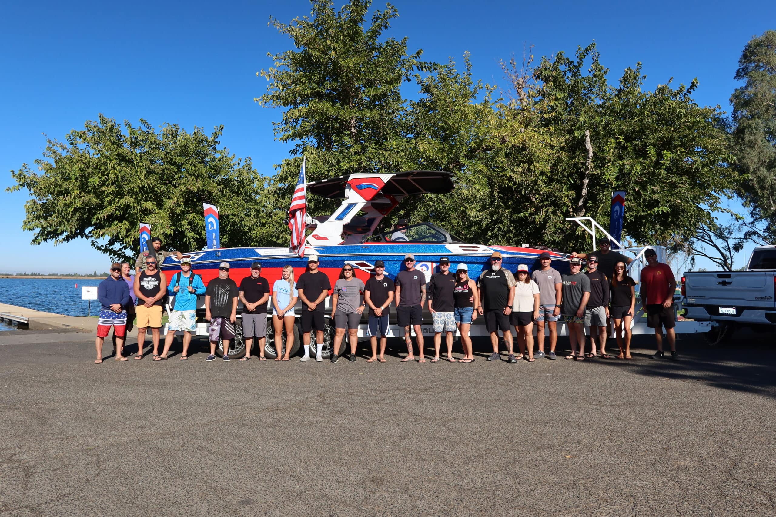 A group of people stands in a line on pavement near a large boat with flags, parked by trees and water on a sunny day.
