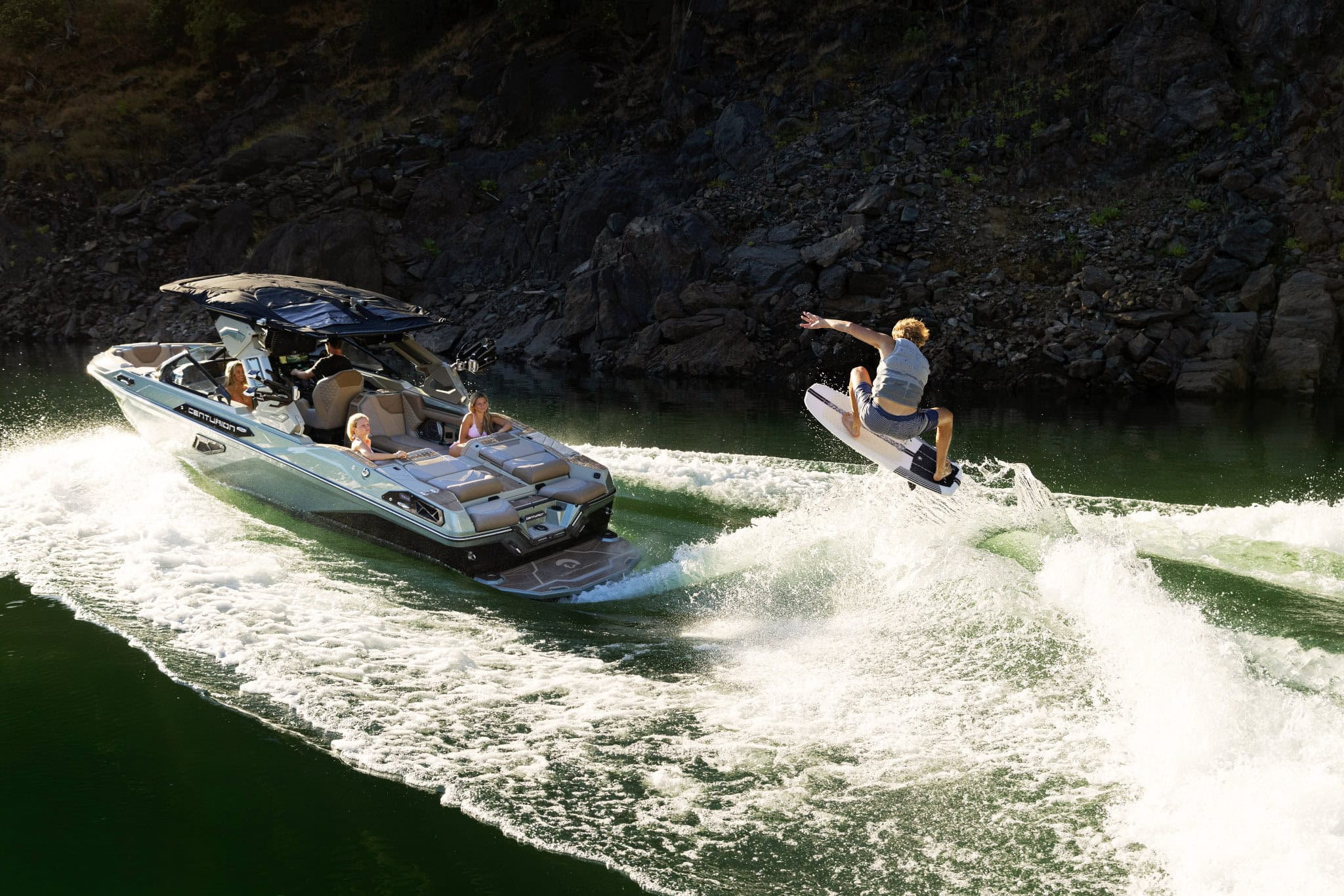 A person rides a wakeboard, catching air behind a speedboat on a green lake, while three people watch from the boat. Rocky shoreline is visible in the background.