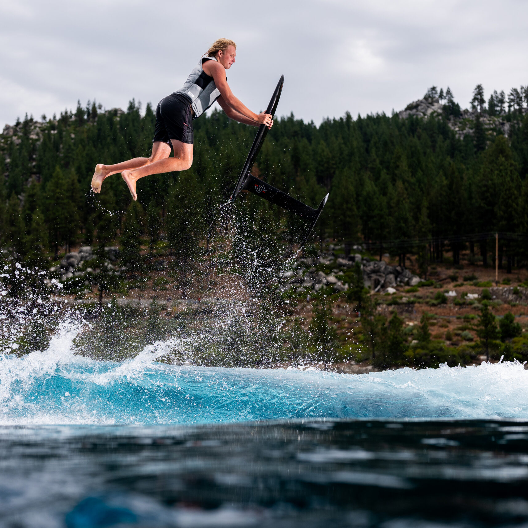 A person in a life jacket rides a hydrofoil board, lifted above the water, with trees and hills in the background under a cloudy sky—perhaps channeling the adventurous spirit of Kendall Brabender.