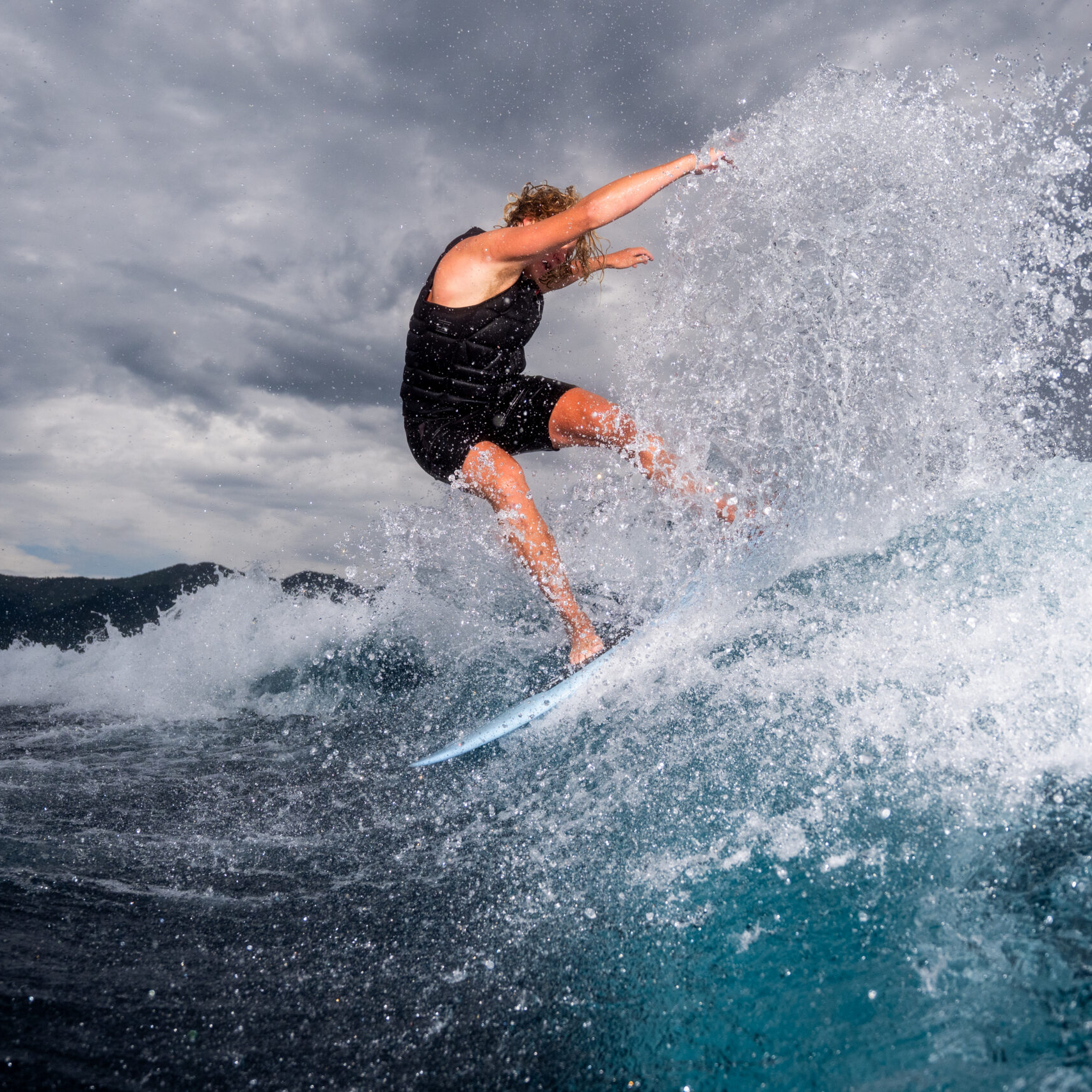 Person in black clothing rides a wave on a surfboard, water splashing around them with a cloudy sky in the background—capturing the thrill of the moment just like Kendall Brabender.