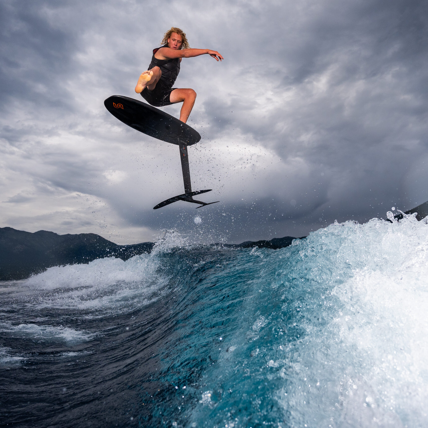 Kendall Brabender rides a hydrofoil surfboard, jumping above a wave with mountains and a cloudy sky in the background.