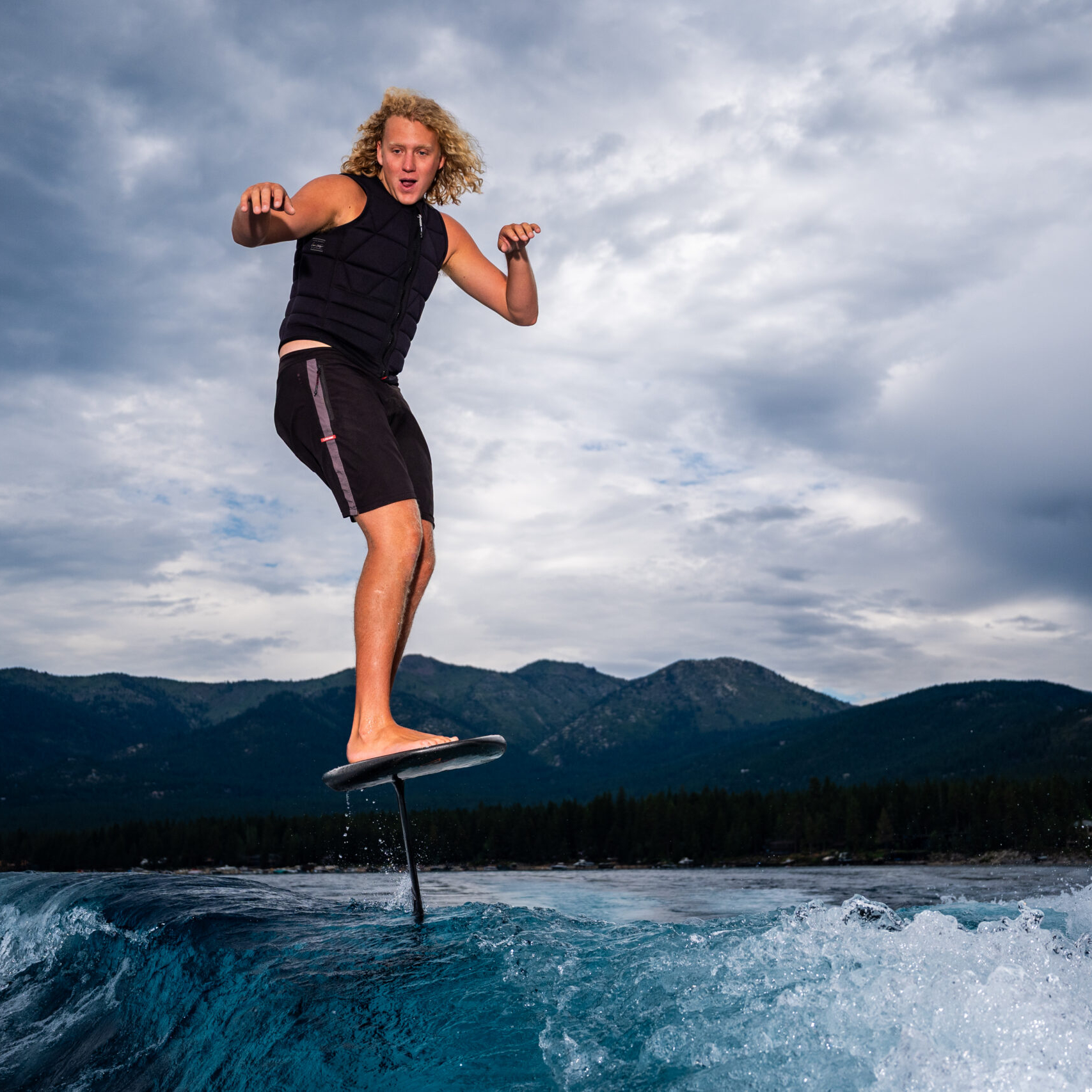 Kendall Brabender, with blonde curly hair, rides a hydrofoil surfboard above the water, wearing a black vest and shorts, with mountains and a cloudy sky in the background.