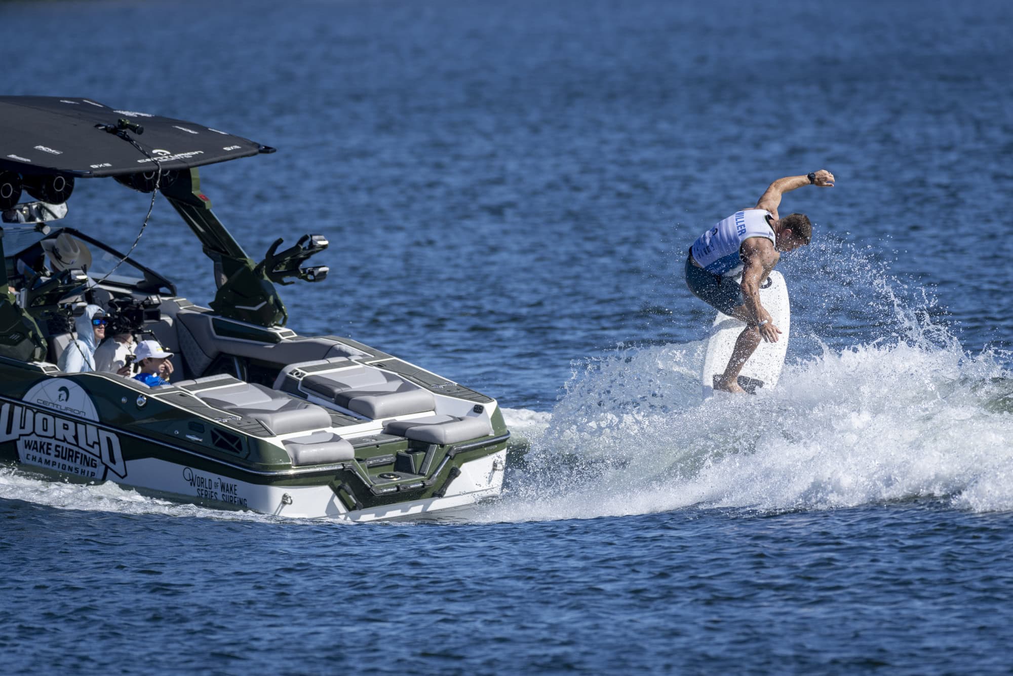 A person wake surfs behind a boat labeled “World Wake Surfing” on a sunny day, with two people seated inside the boat.