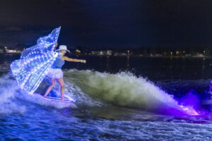 Person wakesurfs at night wearing illuminated wings and a white hat, with bright lights reflecting on the water and a dark shoreline in the background.