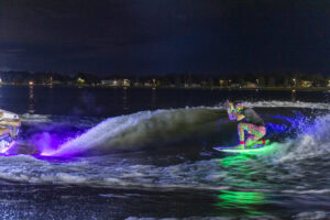 A person rides a neon-lit wakeboard behind a boat at night, creating a colorful glow on the water with lights reflecting in the background.