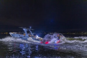 A person wakeboards behind a boat with LED lights at night. The boat displays sponsor logos, and water is illuminated by colorful lights.