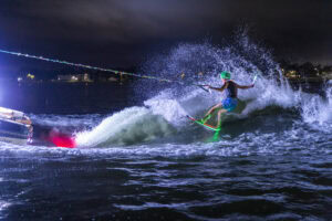 A person wearing a helmet is wakeboarding at night, holding a tow rope behind a boat with colorful lights, water splashing around them.