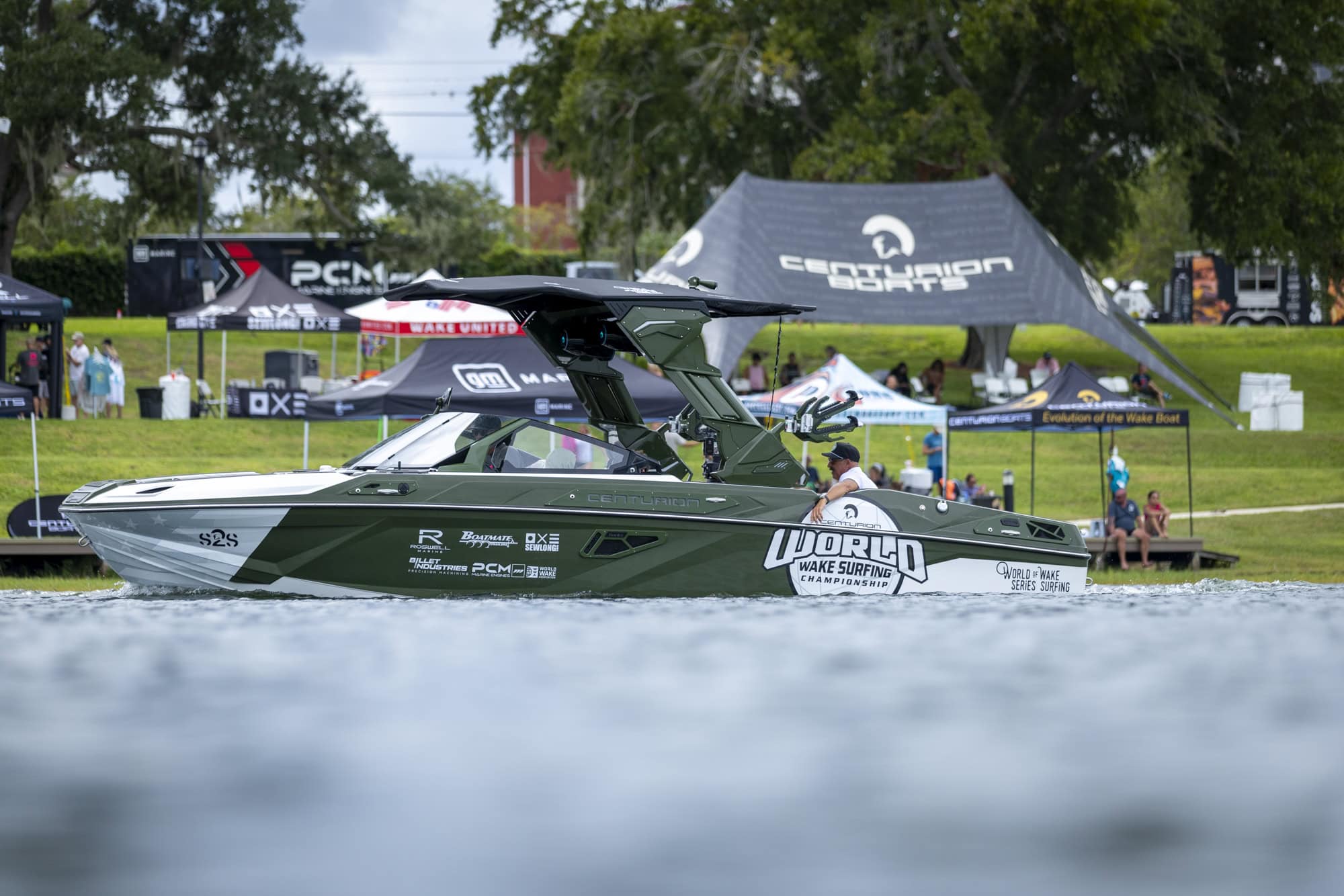 A green and white wakeboarding boat with “World Wake Surfing” decals floats on water near event tents and spectators on a grassy bank.