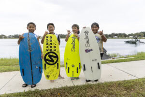 Four children stand on a sidewalk near a lake, each holding a wakesurf board and making a shaka hand gesture, with a boat and trees visible in the background.